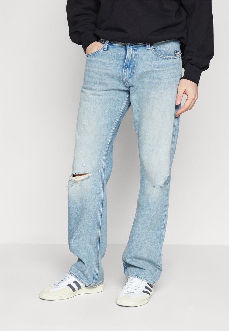Jeans de mezclilla azul claro con un corte relajado, que presentan un desgarro en la rodilla izquierda. Combinados con zapatillas deportivas blancas con franjas negras.