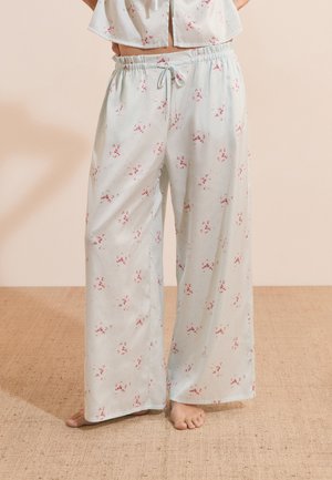 Pantalon de pyjama blanc ample avec petit motif floral rose et taille à cordon, porté pieds nus sur un sol moquetté beige.