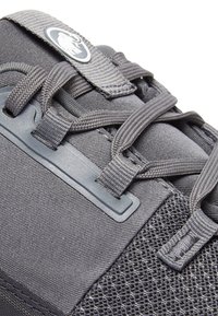 Grå idrottssko med en strukturerad ovandel, nät detaljer, platta snören och en logotyp på en etikett. Har en vadderad tunga och en stabil sula.