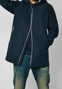 Námořnická voděodolná bunda s kapucí, vybavená celopropínacím zipem a kontrastním světle šedým lemem, kombinovaná s ležérními džínovými kalhotami.