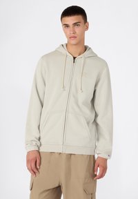 Sudadera color beige claro con cremallera frontal, capucha con cordón y bolsillos tipo canguro. Presenta puños elásticos y un pequeño logo en el pecho.
