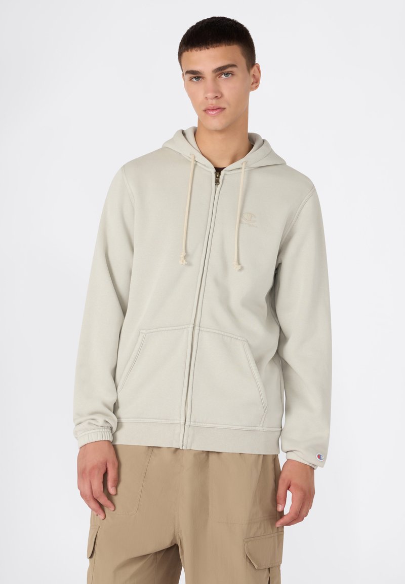 Sudadera color beige claro con cremallera frontal, capucha con cordón y bolsillos tipo canguro. Presenta puños elásticos y un pequeño logo en el pecho.