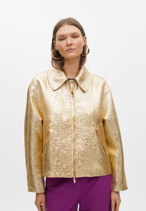 Veste en métal doré avec une surface texturée, grand col, fermeture éclair et poches latérales. Portée avec un pantalon violet.