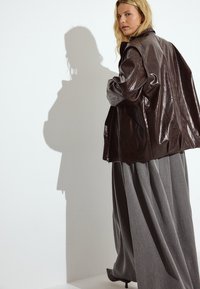 Braune Lederjacke mit glatter Oberfläche, kombiniert mit einem fließenden grauen Faltenrock. Der Hintergrund ist weiß, was einen Kontrast zum Outfit schafft.