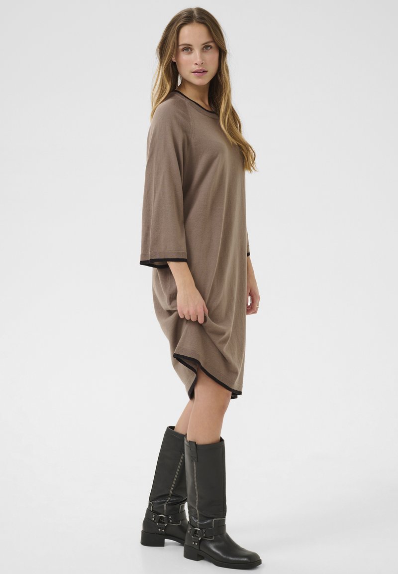 Braun oversized Kleid mit drei Viertel-Ärmeln, versehen mit schwarzem Besatz. Getragen mit knielangen schwarzen Lederstiefeln. Glatte Textur und entspannte Passform.