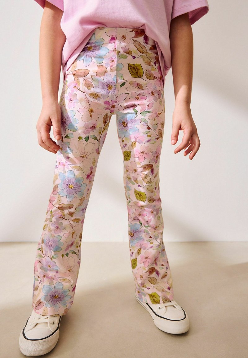 Barn iført pastelblomstrede bukser, hvide sneakers og en lyserød skjorte, stående mod en ensfarvet baggrund.