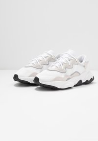 Zapatillas deportivas blancas con parte superior de malla y ante, acentos de diseño en zigzag, punta redonda, cordones blancos y suela de goma negra.