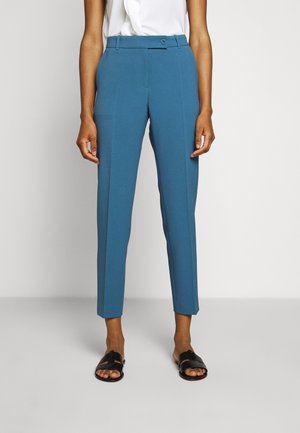 Femme portant un pantalon bleu taille haute, slim, coupé au-dessus de la cheville avec un pli sur le devant, associé à un haut blanc et des sandales noires à enfiler.