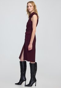 Abito senza maniche borgogna con colletto, chiusura frontale a bottoni, due tasche e uno spacco laterale, abbinato a stivali neri al ginocchio.