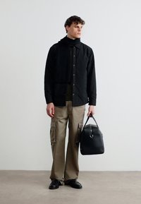 Άνδρας που φορά μαύρο μπουφάν και κασκόλ, μπεζ παντελόνι καρό, μαύρα παπούτσια, κρατώντας μαύρη δερμάτινη τσάντα duffle, στέκεται μπροστά σε μια απλή λευκή τοίχο.