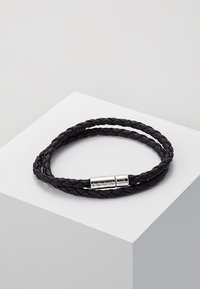 Svart flettet lær armbånd med en lukking i sølvfarge merket med "ICON." Designet er stilrent og vikler seg to ganger rundt håndleddet.