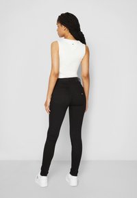 Mulher jovem com cabelo trançado, vestindo uma blusa branca sem mangas, jeans justos pretos e ténis brancos, de pé com o corpo de costas em um fundo simples.