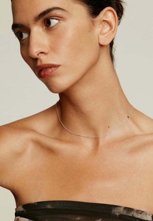 Collar de cadena de plata con finos eslabones redondos y una pequeña cuenta de acento. La modelo lo lleva con una blusa sin hombros.