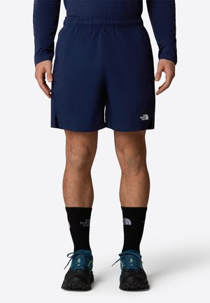 Mann trägt marineblaue Sportshorts, schwarze Crew-Socken und blaue Laufschuhe und steht vor einem schlichten Hintergrund.