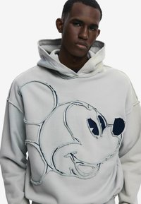 Sudadera blanca con capucha que presenta bordes deshilachados y un diseño de Mickey Mouse cosido. La tela parece suave con un ajuste casual.