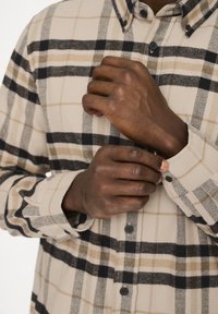 Beige- und schwarze Karoshemd mit kontrastierenden sandfarbenen Akzenten, ausgestattet mit einem Standardkragen und schwarzen Knopfverschlüssen, präsentiert auf verschränkten Armen.