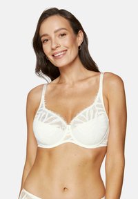 Biały koronkowy biustonosz z przezroczystymi panelami, kwiatowymi wzorami, falbankami i regulowanymi ramiączkami. Modelka prezentuje widok z przodu.