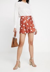 Oranžové šortky s puntíky, vysokým pasem a páskem, v kombinaci s bílou blůzou s dlouhým rukávem a zlatými sandály na blokovém podpatku. Hnědá kabelka.