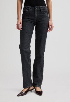 Kobieta w czarnych dżinsach z wysokim stanem i prostymi nogawkami oraz w ciemnobrązowych butach na niskim obcasie z ostrym noskiem, stojąca na jednolitym tle.