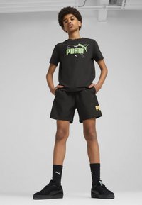 Camiseta de algodón negra con logo verde, combinada con pantalones cortos negros que tienen texto amarillo. Zapatillas y calcetines negros completan el conjunto.