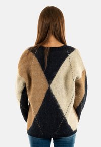 Persona con cabello largo que lleva un suéter de patrón argyle en marrón, negro y crèma, de pie mirando hacia atrás frente a un fondo sencillo.