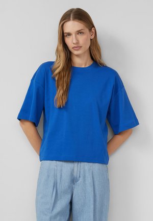 Kobieta o długich blond włosach, ubrana w jasnoniebieską, oversize'ową koszulkę i jasnoniebieskie jeansy z wysokim stanem, stojąca na szarym tle.