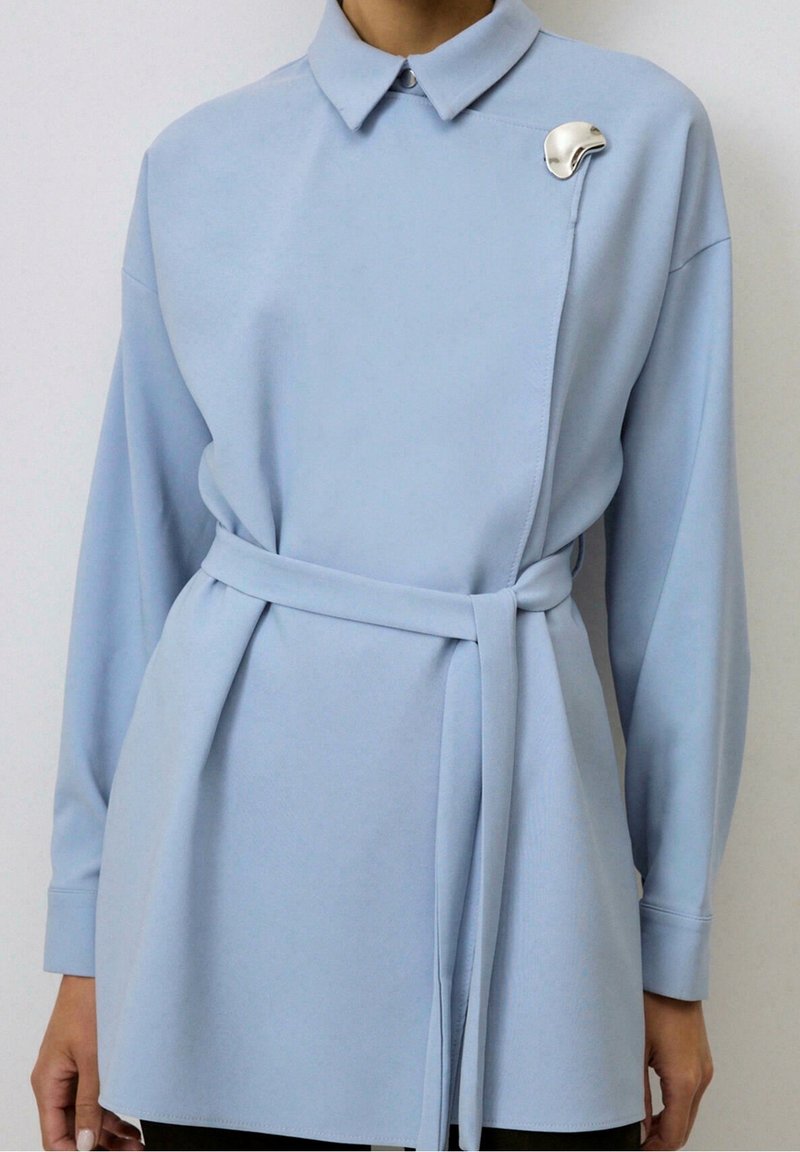 Personne portant une tunique bleu clair à manches longues, ceinturée, avec un col et une broche abstraite argentée sur l'épaule gauche.
