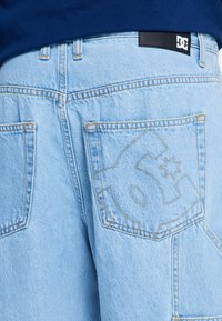 Jasnoniebieskie jeansy z denimu z naszytym logo DC na tylnej kieszeni oraz czarną naszywką z logo na talii, noszone z ciemnoniebieską bluzką.