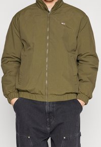 Giacca bomber verde oliva con zip frontale, polsini e vita elasticizzati. Include un piccolo logo sulla parte superiore sinistra del petto.