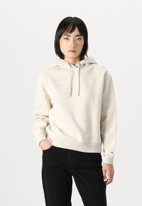 Un hoodie couleur crème avec des cordons de serrage et une texture travaillée, arborant un logo dans une teinte plus claire. Associé à un pantalon noir.