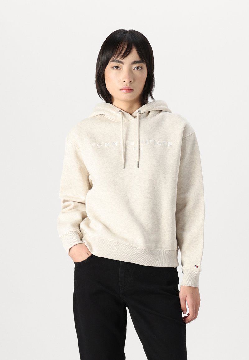 Un hoodie couleur crème avec des cordons de serrage et une texture travaillée, arborant un logo dans une teinte plus claire. Associé à un pantalon noir.