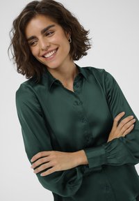Dark green satin blouse with a button-up front, pointed collar, and gathered cuffs. Smooth texture with subtle sheen.