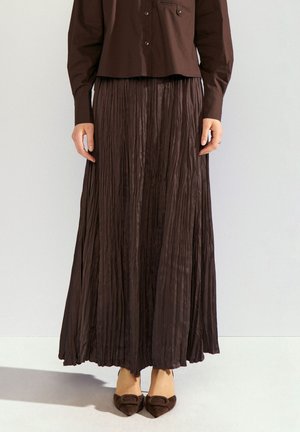 Femme portant une longue jupe plissée brun foncé, une chemise brun foncé boutonnée et des chaussures plates assorties à bout pointu avec détail de boucle.