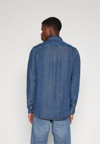 Chemise en denim bleu moyen, dotée d'un col, de poignets à boutons et d'une coupe droite, avec des détails de couture le long des bords.