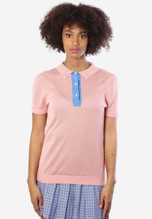 Junge Frau mit lockigem Haar, die ein pinkes Kurzarmshirt mit einer blauen Knopfleiste und einen blau-pinken karierten Rock trägt.