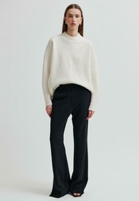 Kobieta z długimi włosami, ubrana w luźny biały sweter, czarne szerokie spodnie i czarne buty na odkrytych palcach, stojąca na jednolitym tle.