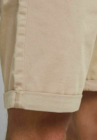 Pantalones cortos beige hechos de una tela tejida, con un dobladillo enrollado y costuras laterales. La textura es suave, y el diseño es simple y funcional.