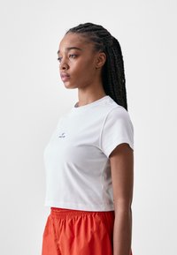 White cropped t-shirt with short sleeves and a round neckline, featuring a small blue logo on the chest. Model paired with orange shorts.