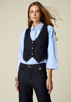 Femme aux longs cheveux portant une chemise bleu clair avec des détails en perles sur le col, un gilet en denim foncé avec des boutons, un pantalon en denim foncé assorti et des boucles d’oreilles argentées.