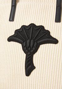 Écusson en cuir noir en forme d'éventail avec des détails texturés sur un fond en tissu beige tissé.