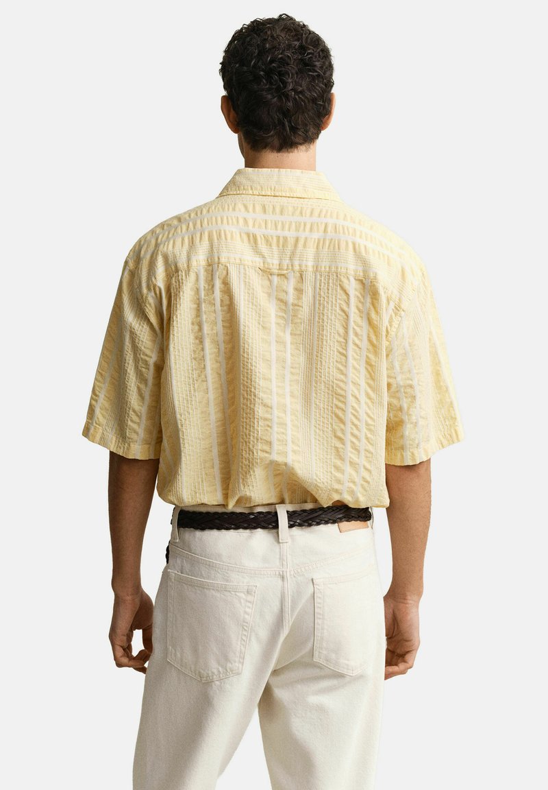 Ein Mann von hinten gesehen, der ein hellgelbes, kurzärmeliges, gestreiftes Hemd trägt, das in weiße Hosen mit einem dunklen, gewebten Gürtel gesteckt ist.