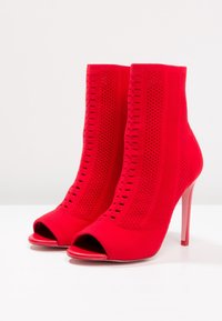 Bottines rouges en tricot, à talons aiguilles hauts et bouts ouverts, avec des détails perforés sur les côtés. Texture lisse dans l'ensemble.