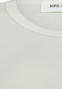 Fehér, bordázott felső, kerek nyakkivágással és vékony textúrával. A "SOFIE" felirat látható a gallér közelében.