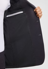 Zwarte blazer binnenkant met rode stiksel, met één witte ritszak en een over het algemeen gladde, strakke stofstructuur.