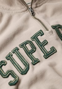 Sweat-shirt zippé beige avec des lettres brodées en vert foncé épelant "SUPER". Le tissu a une texture douce et des détails de couture visibles.