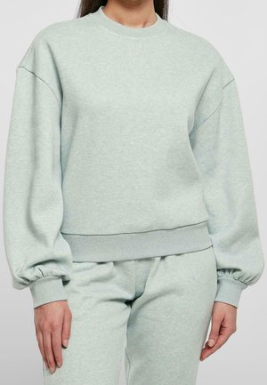 Femme portant un sweat-shirt vert clair oversize avec des manches ballon et un pantalon assorti sur un fond uni.