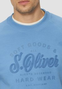 Felpa azzurra chiara con un design del logo testurizzato in blu scuro, con colletto a costine e testo stampato in modo sottile. Materiale misto cotone.
