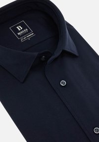 Námorně modrá košile na knoflíky vyrobená z texturované látky, zdobená rozloženým límcem a dvěma odpovídajícími knoflíky vpředu.