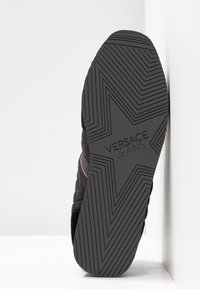 Semelle de basket noire avec un motif texturé en zigzag. Présente l'inscription "VERSACE JEANS" en relief et une forme d'étoile. Le matériau semble être du caoutchouc.