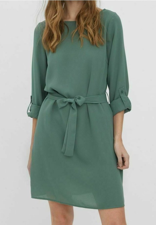Femme portant une robe verte mi-longue à manches longues avec des poignets retroussés et une ceinture en tissu nouée à la taille sur un fond uni.